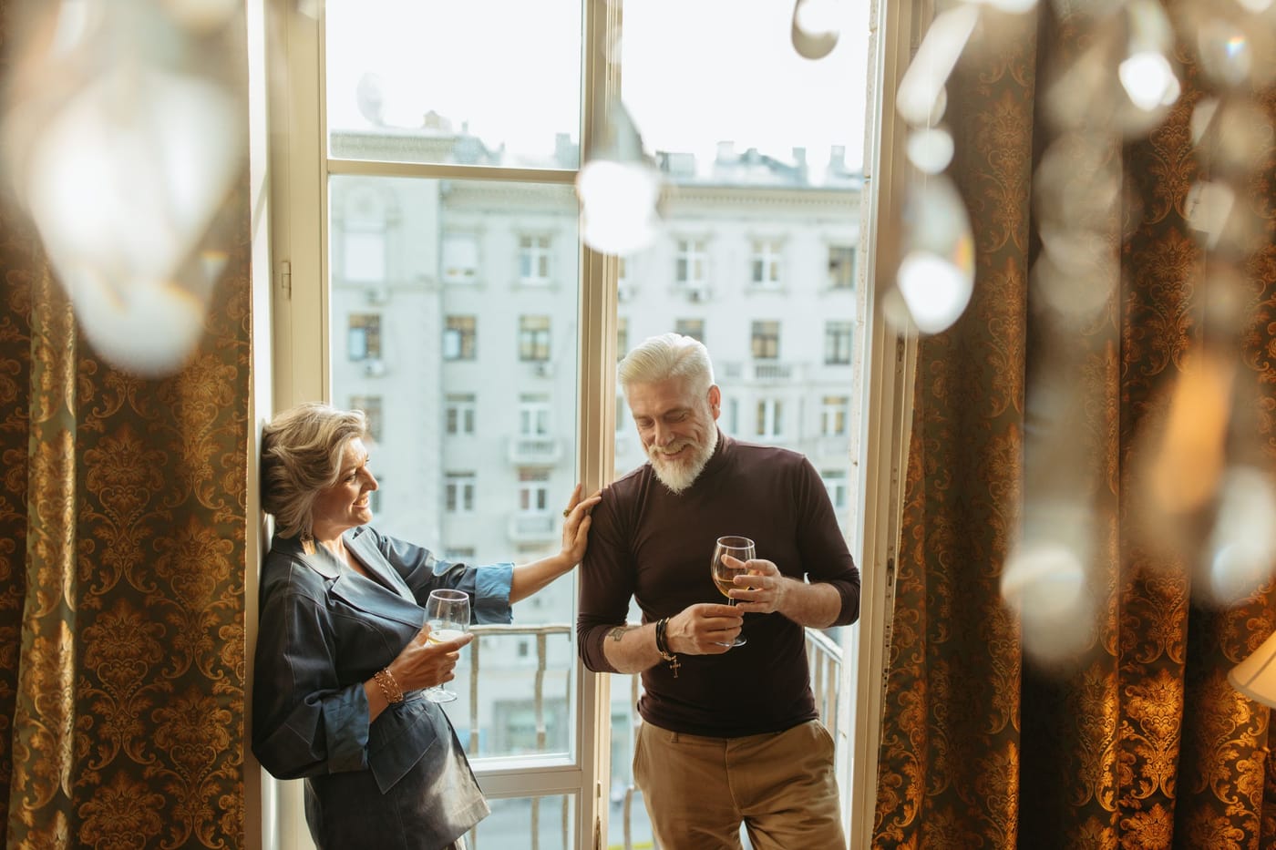Ein Mann mit weißem Bart lacht unter einem Kronleuchter, ein Glas Sekt in der Hand.