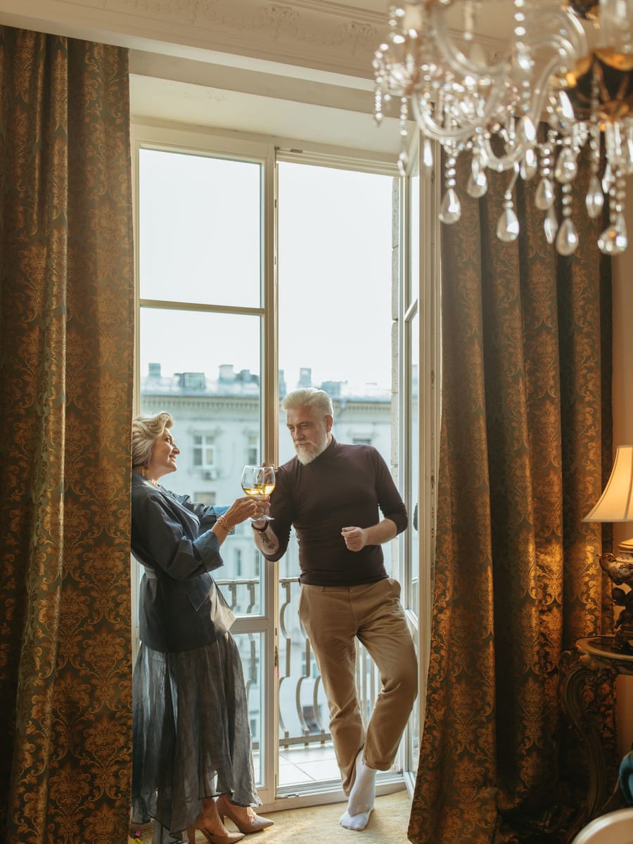 Ein Paar stößt am hohen Fenster eines Altbauzimmers mit Kronleuchter an.